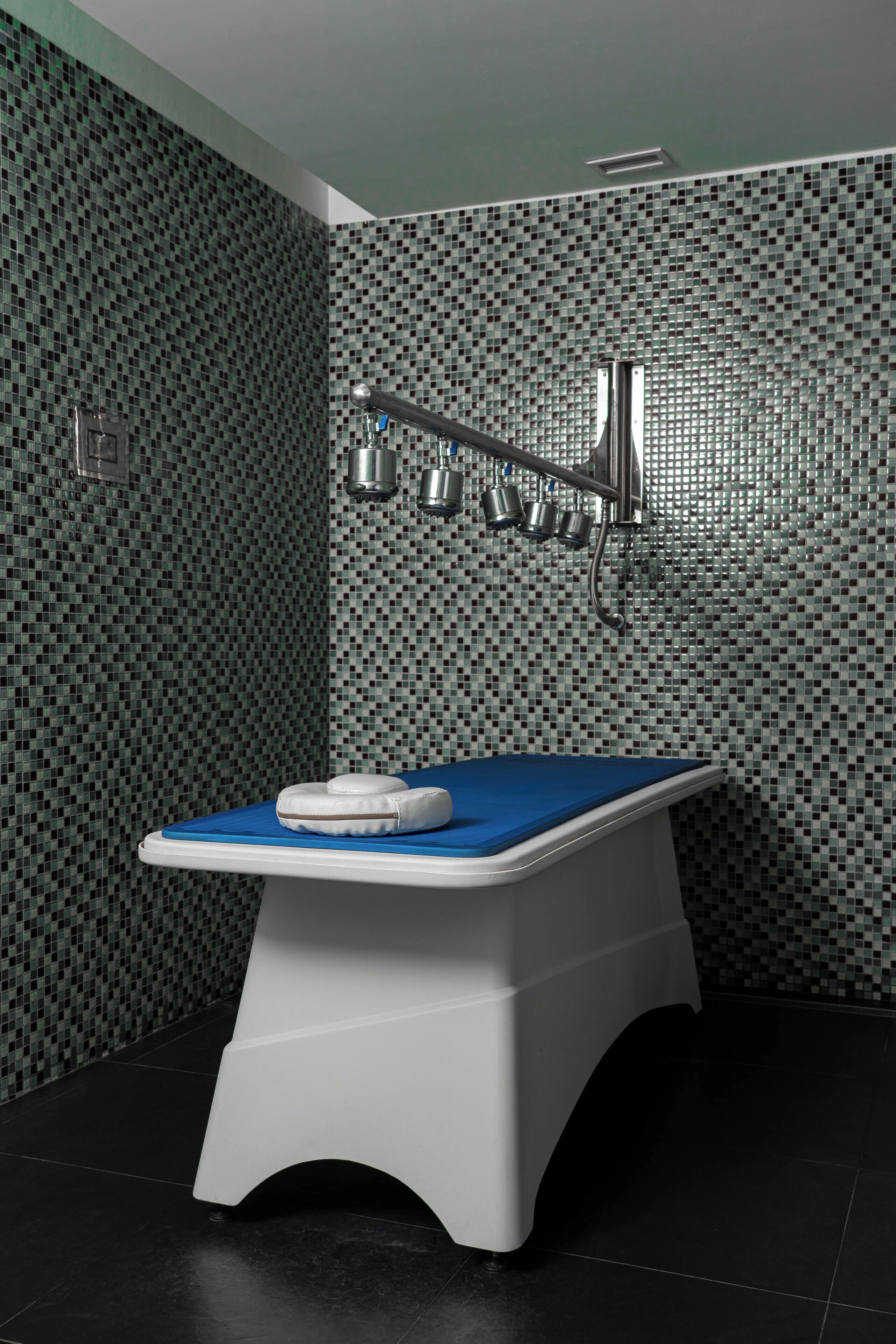 A modern bathroom with a blue-topped examination table, a tiled mosaic wall, and a shower area with a chrome fixture.