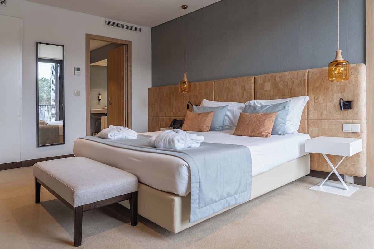 A modern bedroom with a large bed, a bench, a bedside table, and a hanging light fixture.