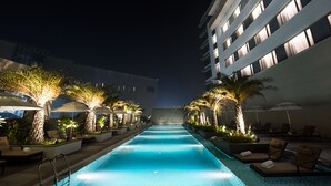 Un área de piscina nocturna con sillones de descanso y sombrillas, rodeada de palmeras y edificios modernos.
