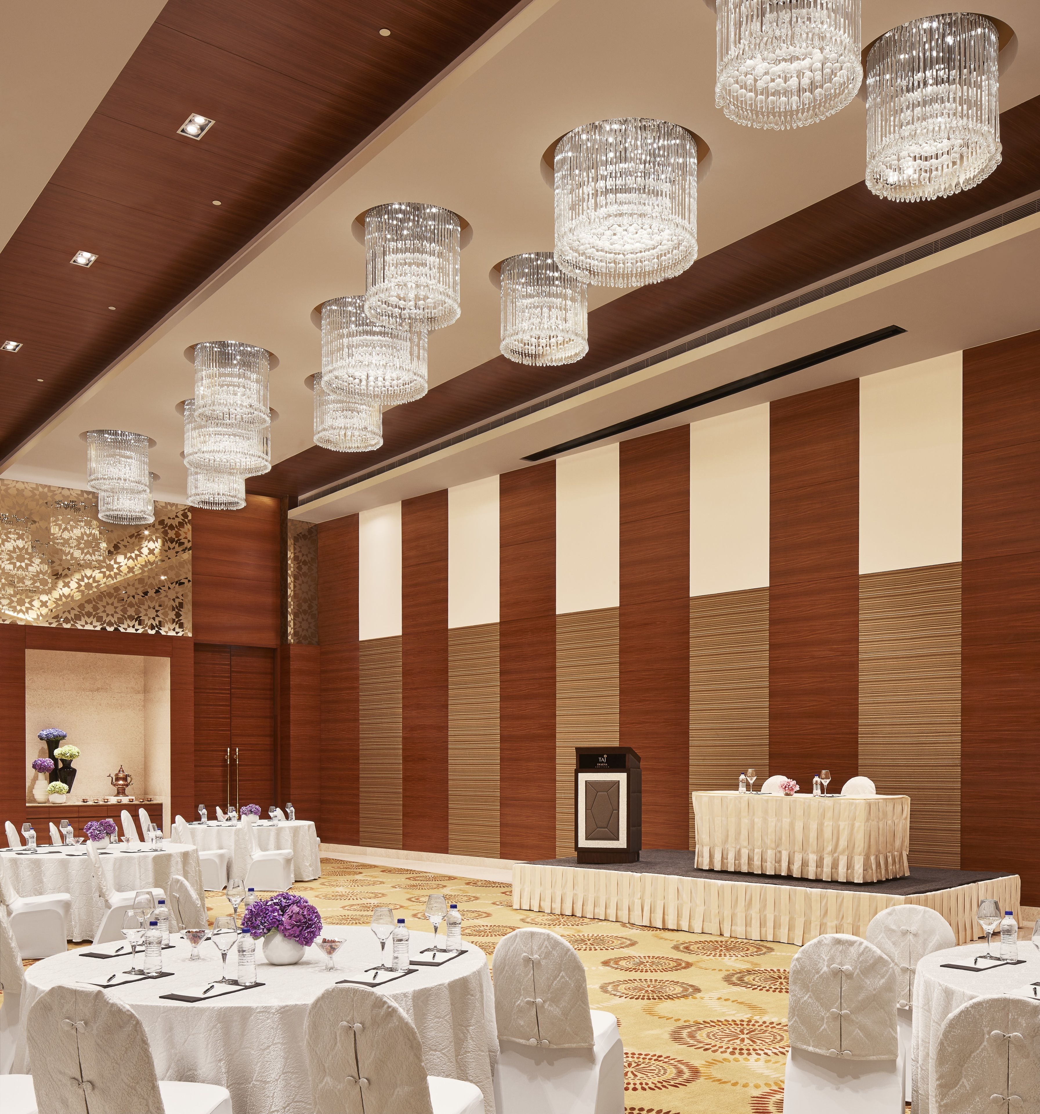 A banquet hall with round tables, white chairs, and chandeliers.