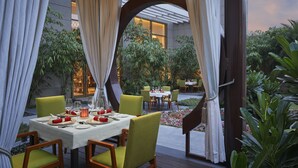 Un área de comedor al aire libre con mesas preparadas para dos, rodeada de follaje y un edificio moderno al fondo.