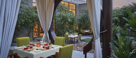 An outdoor dining area with tables set for two, surrounded by greenery and a modern building in the background.