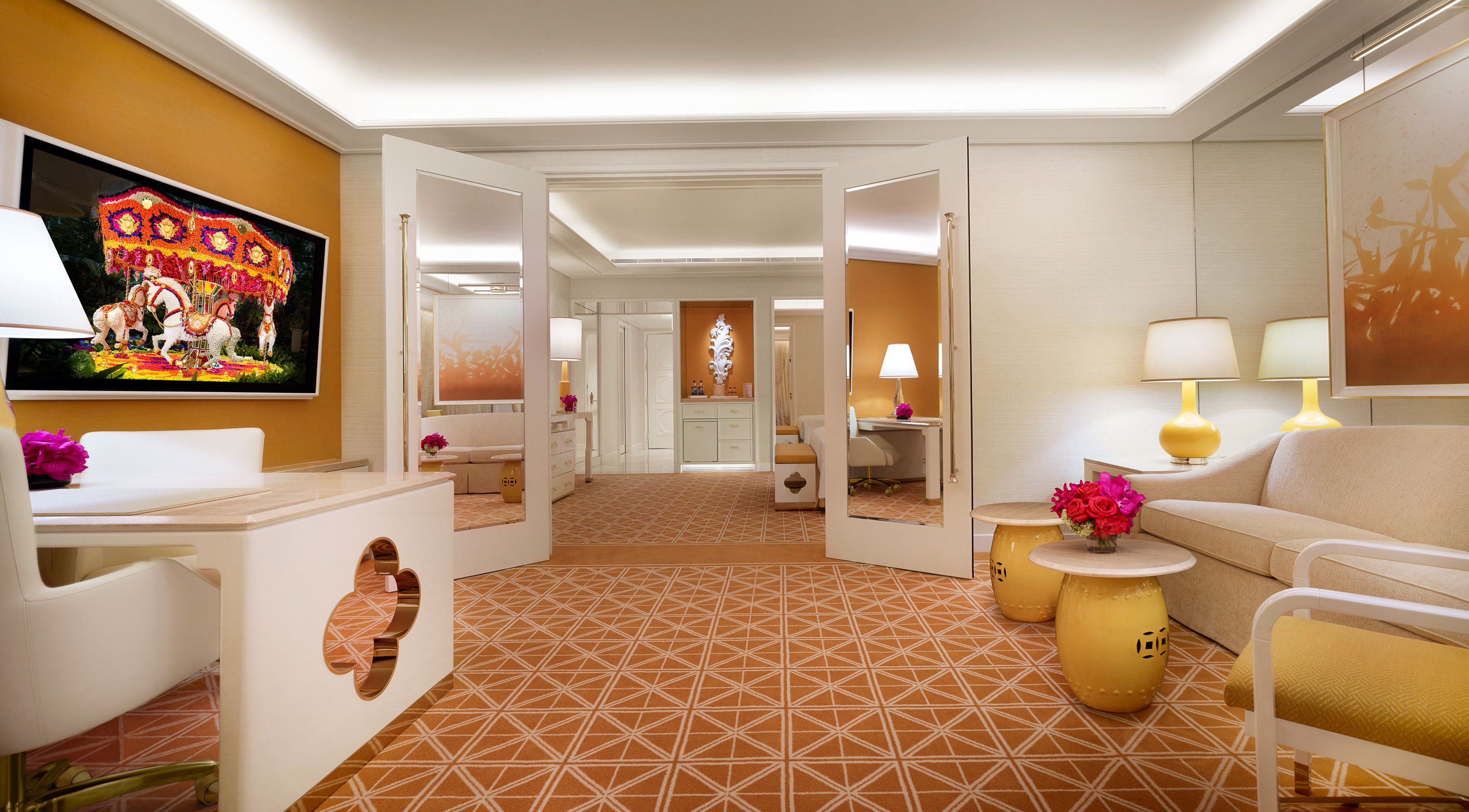 Une chambre d’hôtel avec un sol à motifs géométriques, un canapé, une petite table ronde ornée d’un vase de fleurs, un téléviseur diffusant une scène colorée et un miroir qui reflète la pièce.
