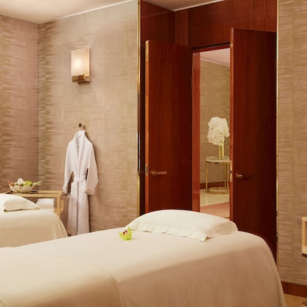 A hotel room with two beds, a wooden wardrobe, a vanity with a mirror, and a small table with towels and toiletries.
