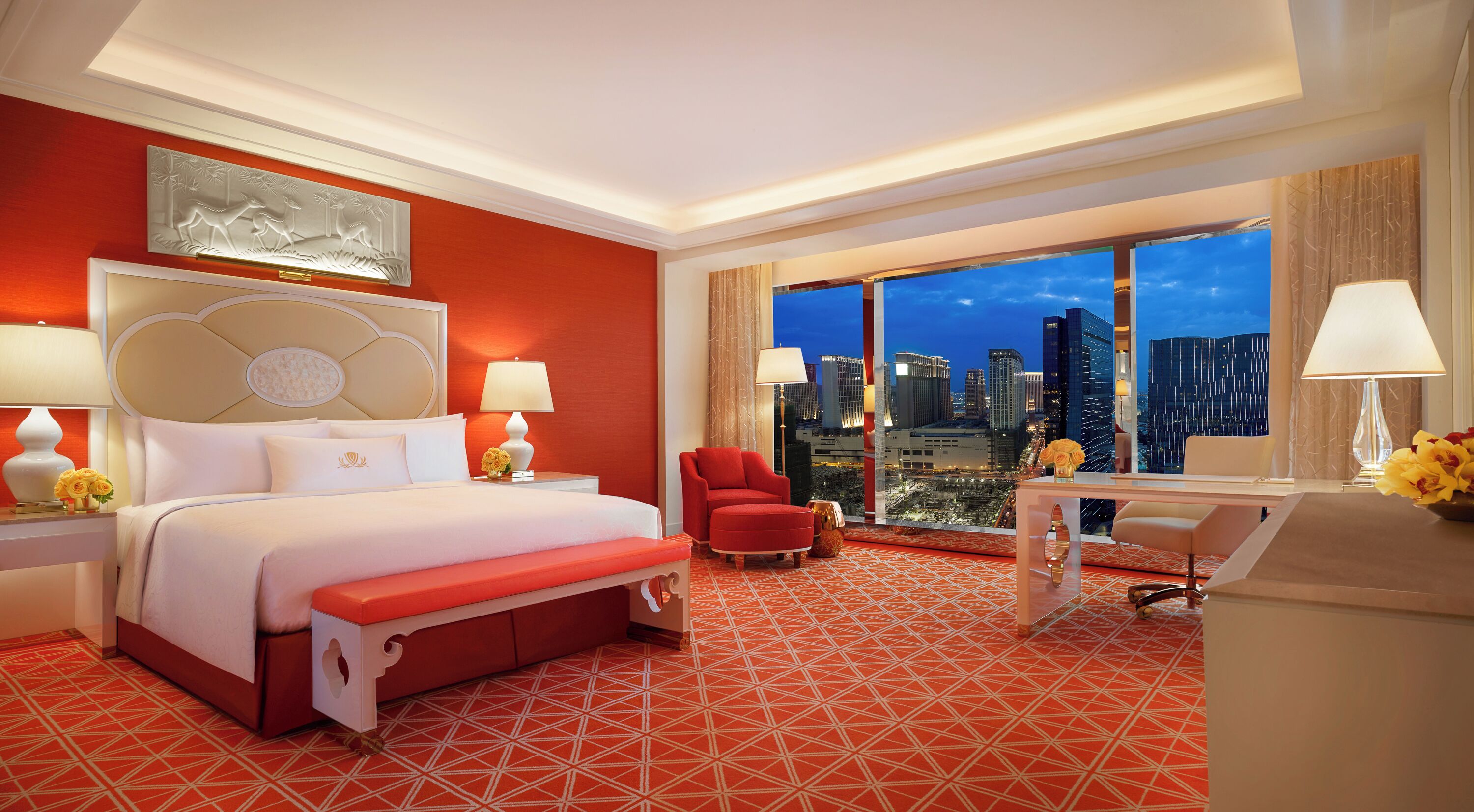 Een hotelkamer met een groot bed, een rode fauteuil, een bureau en uitzicht op de stad 's nachts.