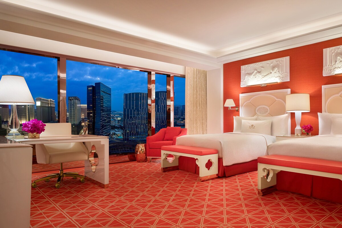 Une chambre d’hôtel avec vue sur la ville, un bureau avec une lampe, une chaise rouge, un lit avec une literie blanche et un tapis rouge à motifs.