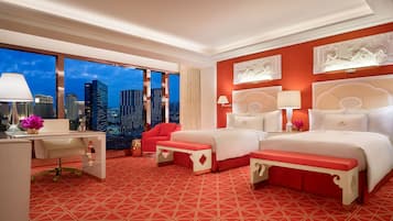 A hotel room with a city view, a desk with a lamp, a red chair, a bed with white bedding, and a red patterned carpet.