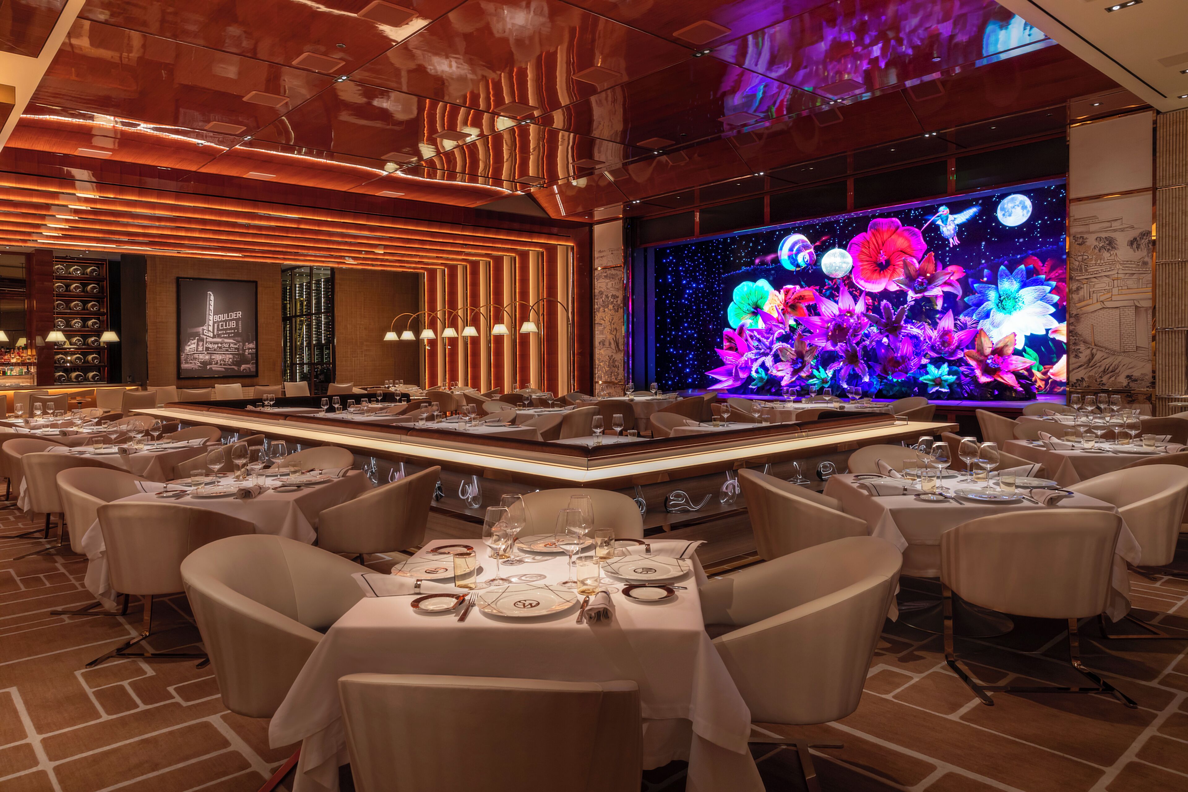 Un restaurante con mesas redondas, manteles blancos y una gran decoración floral iluminada en la pared.