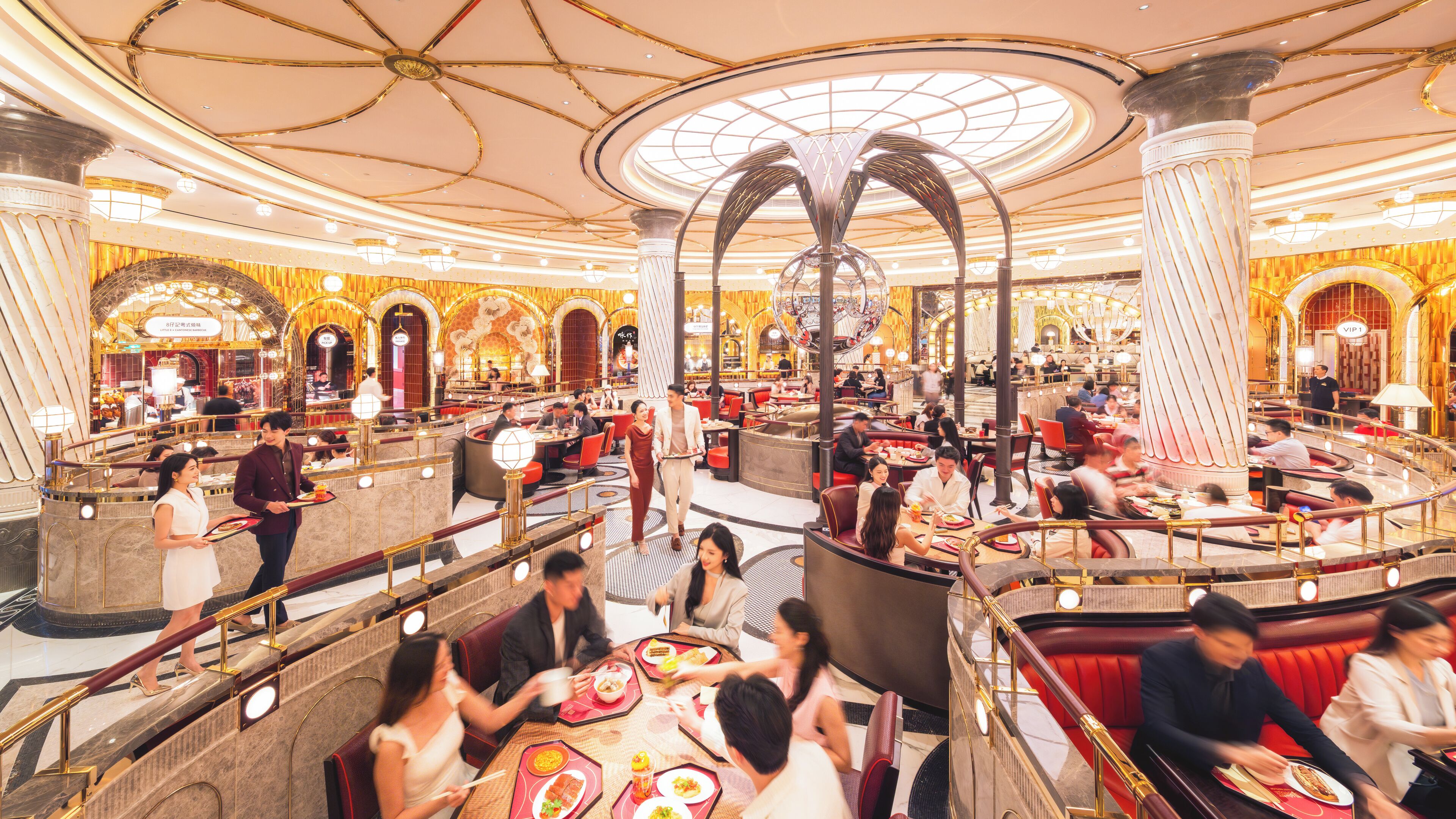 Um restaurante espaçoso, em vários níveis, com arquitetura ornamentada, incluindo uma cúpula central e colunas, e várias áreas de refeições com assentos vermelhos.