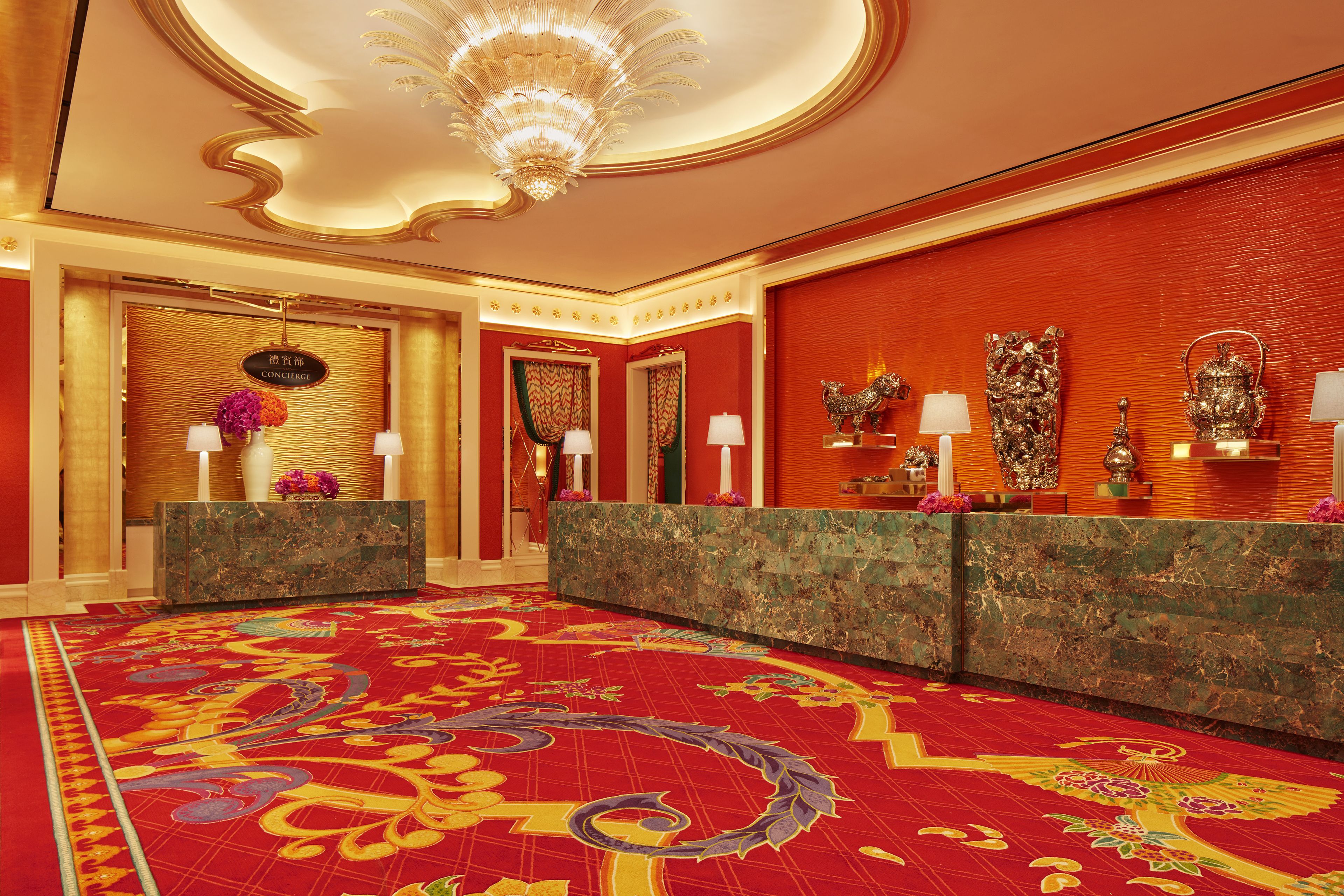 A luxurious lobby with a red carpet, marble reception desk, and ornate wall decorations.
