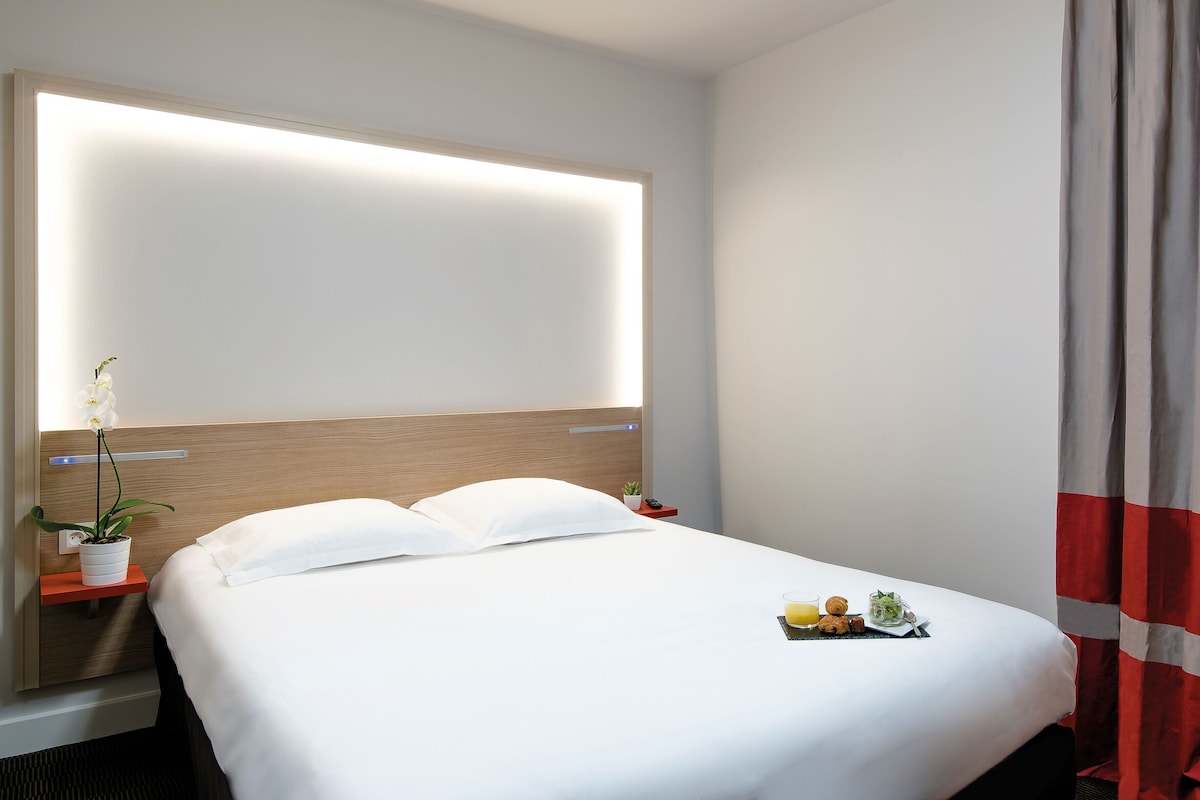 A hotel room with a bed, bedside table, nightstand, a potted plant, and a breakfast tray.