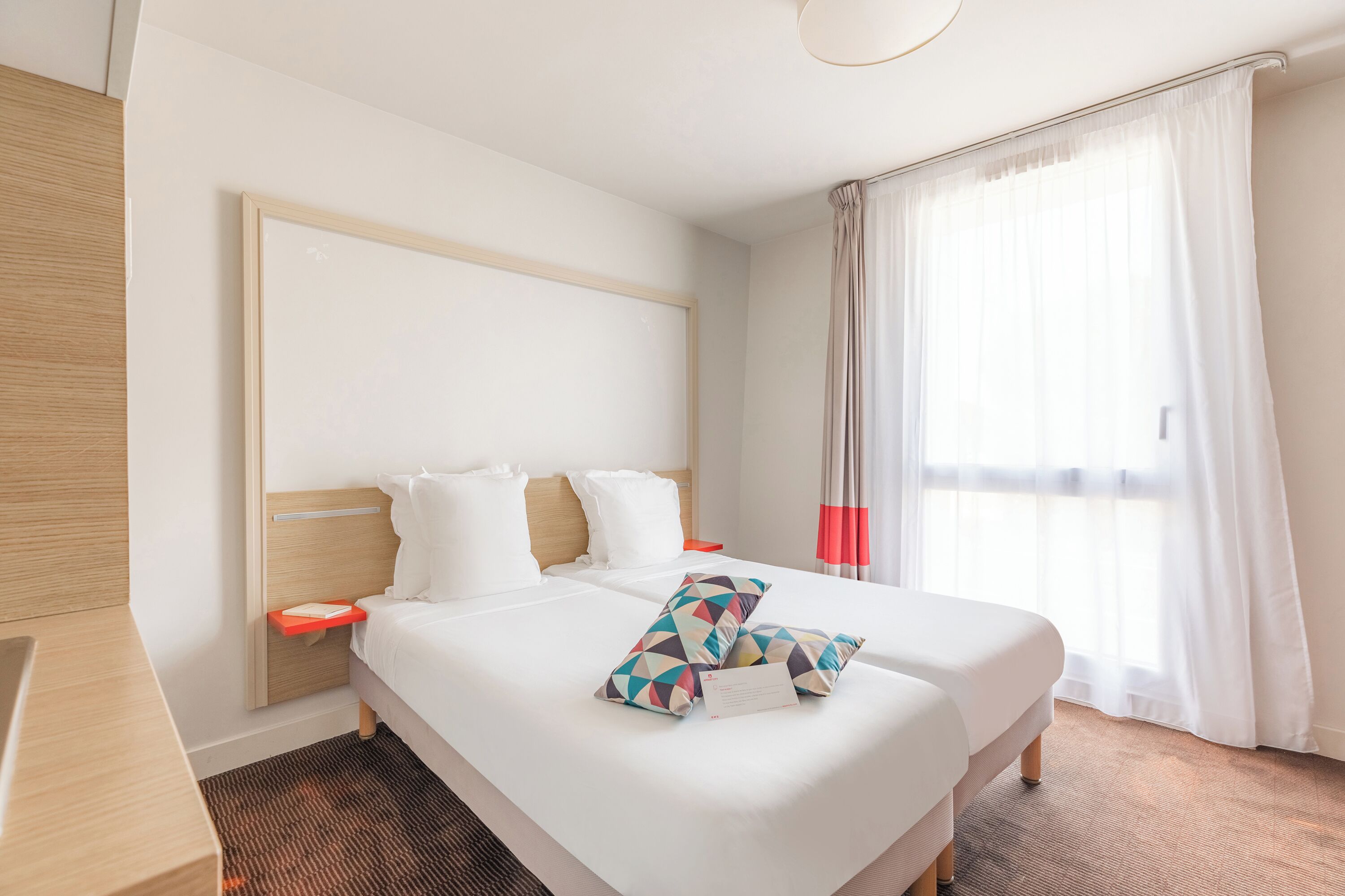 A hotel room with a large bed, a wooden headboard, a bedside table, and a window with sheer curtains.