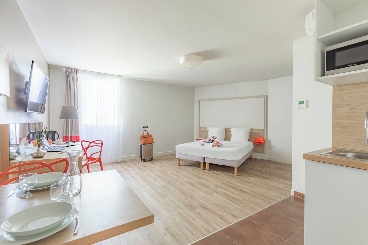 Een moderne hotelkamer met een bed, een bureau met een computer, een eettafel met stoelen en een keukenhoek met een magnetron.