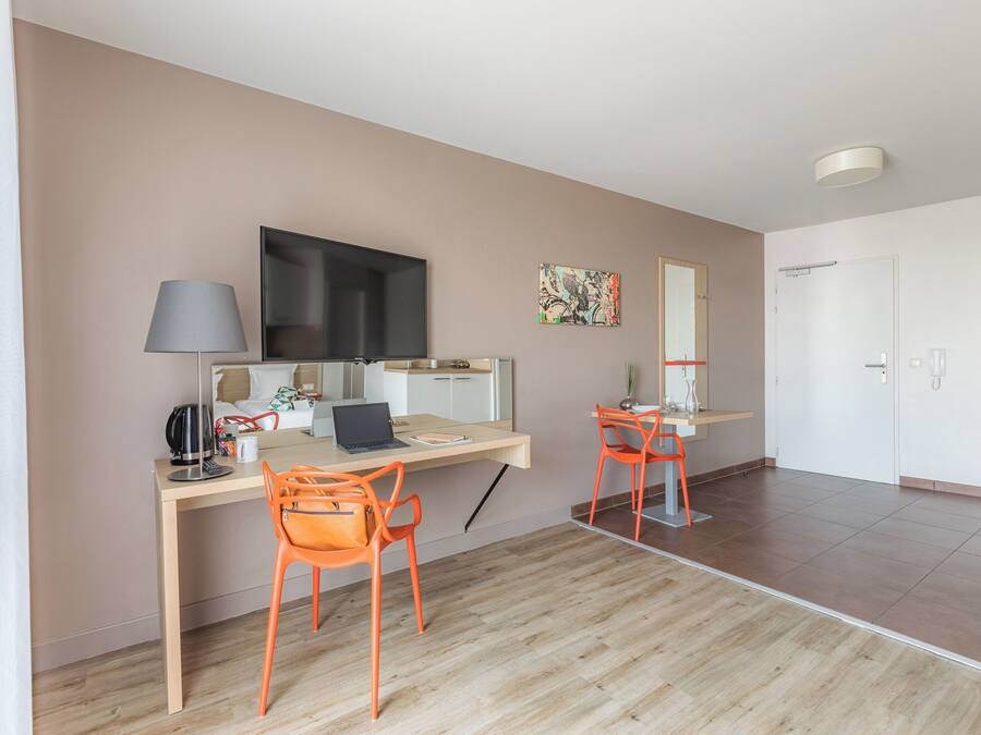 Una habitación moderna con un escritorio, dos sillas naranjas, una computadora portátil, un televisor de pantalla plana, una lámpara y una pequeña planta.