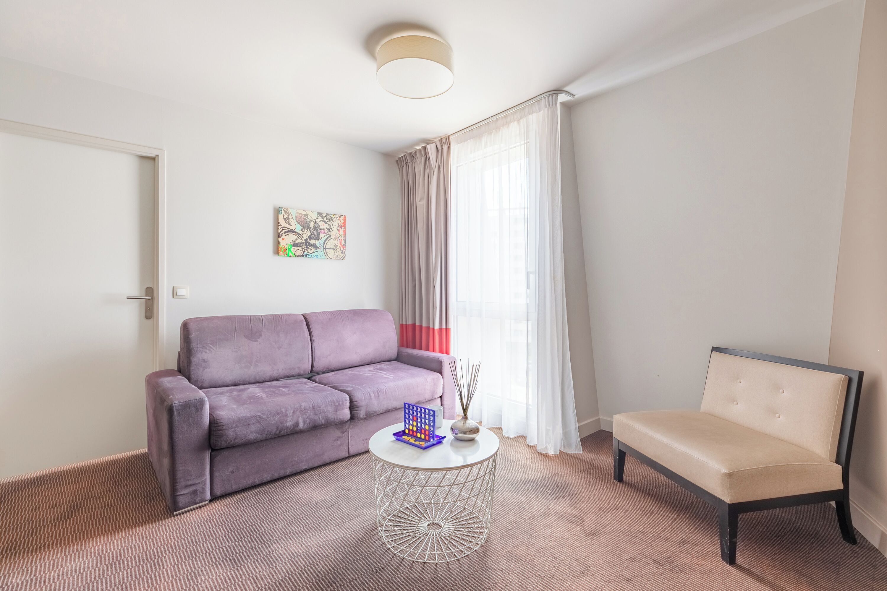 A living room with a purple sofa, a beige armchair, a round coffee table, and a window with sheer curtains.