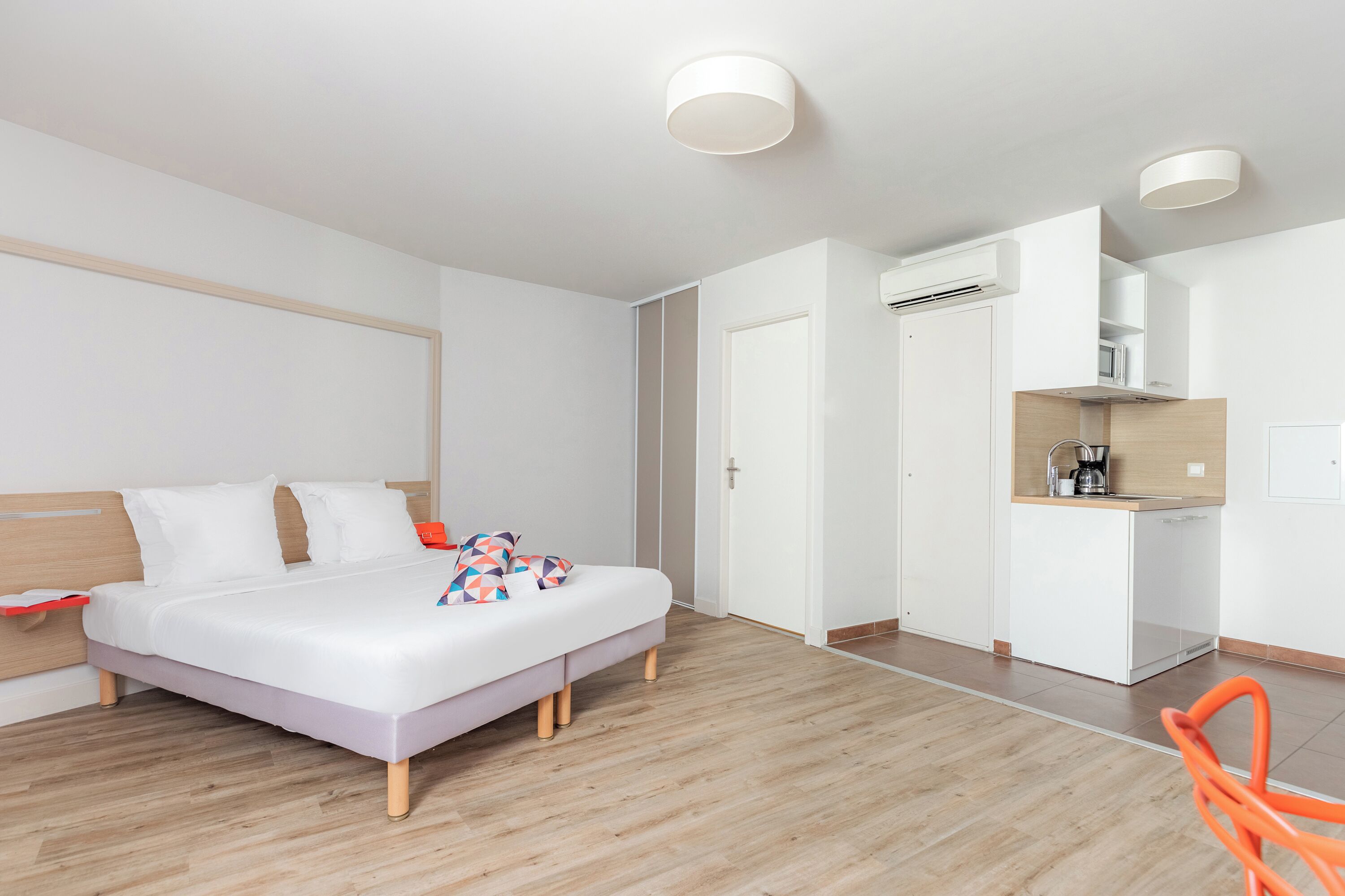 A modern hotel room with a bed, a small kitchenette, and a wooden floor.