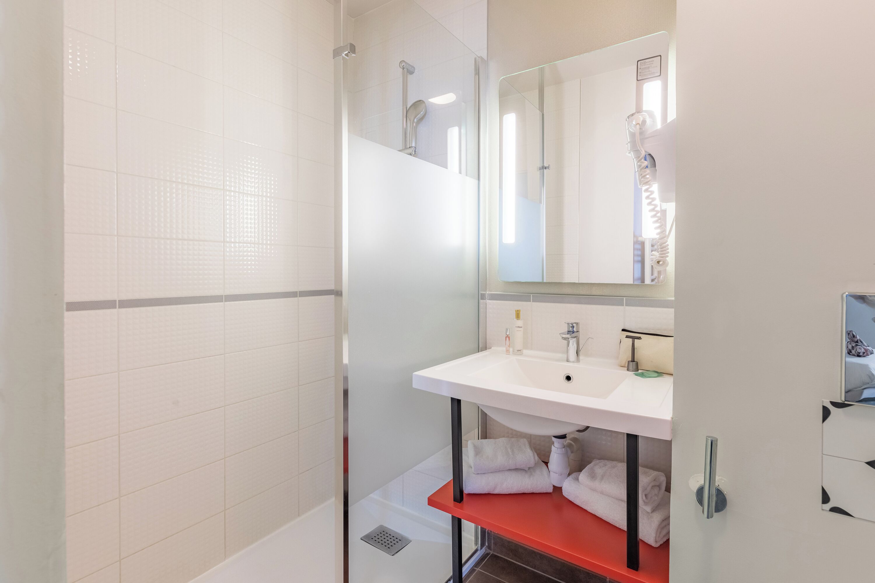 Une salle de bain moderne avec un lavabo blanc, une douche en verre et une étagère rouge où sont posées des serviettes.