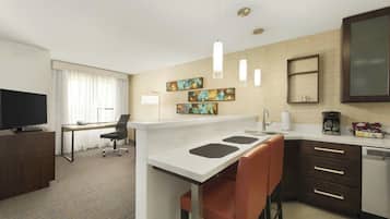 A modern hotel room with a kitchenette, a dining area, a TV, and artwork on the walls.