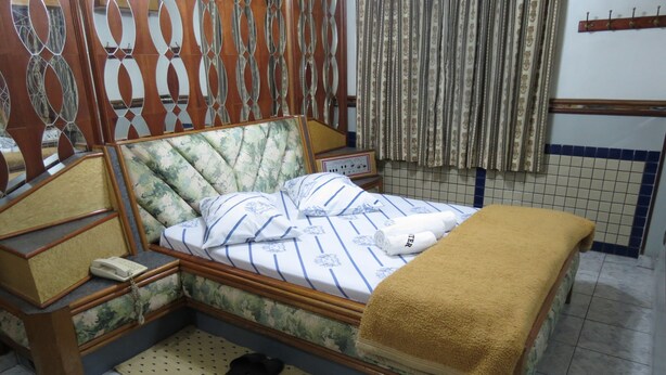 A neatly made bed with a floral headboard, a bedside table with a telephone, and a pair of slippers on a mat. - Hotel PC (São Paulo)