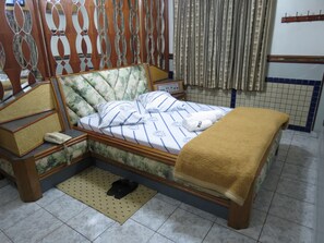 Een netjes opgemaakt bed met een bloemrijk hoofdbord, een nachtkastje met een telefoon, en een paar slippers op een mat.