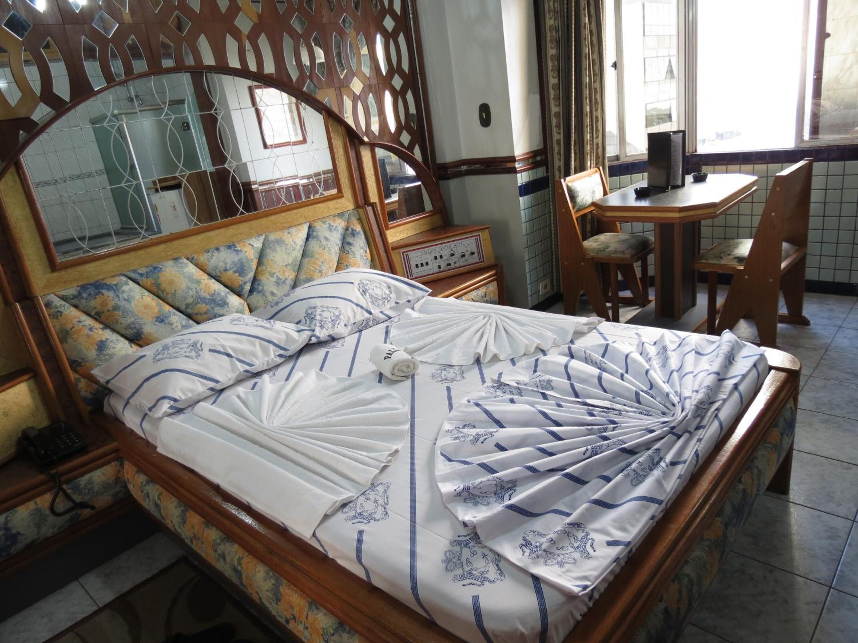 Una cama bien hecha con un cabecero floral, una mesita de noche de madera con una radio y un comedor con mesa y sillas.
