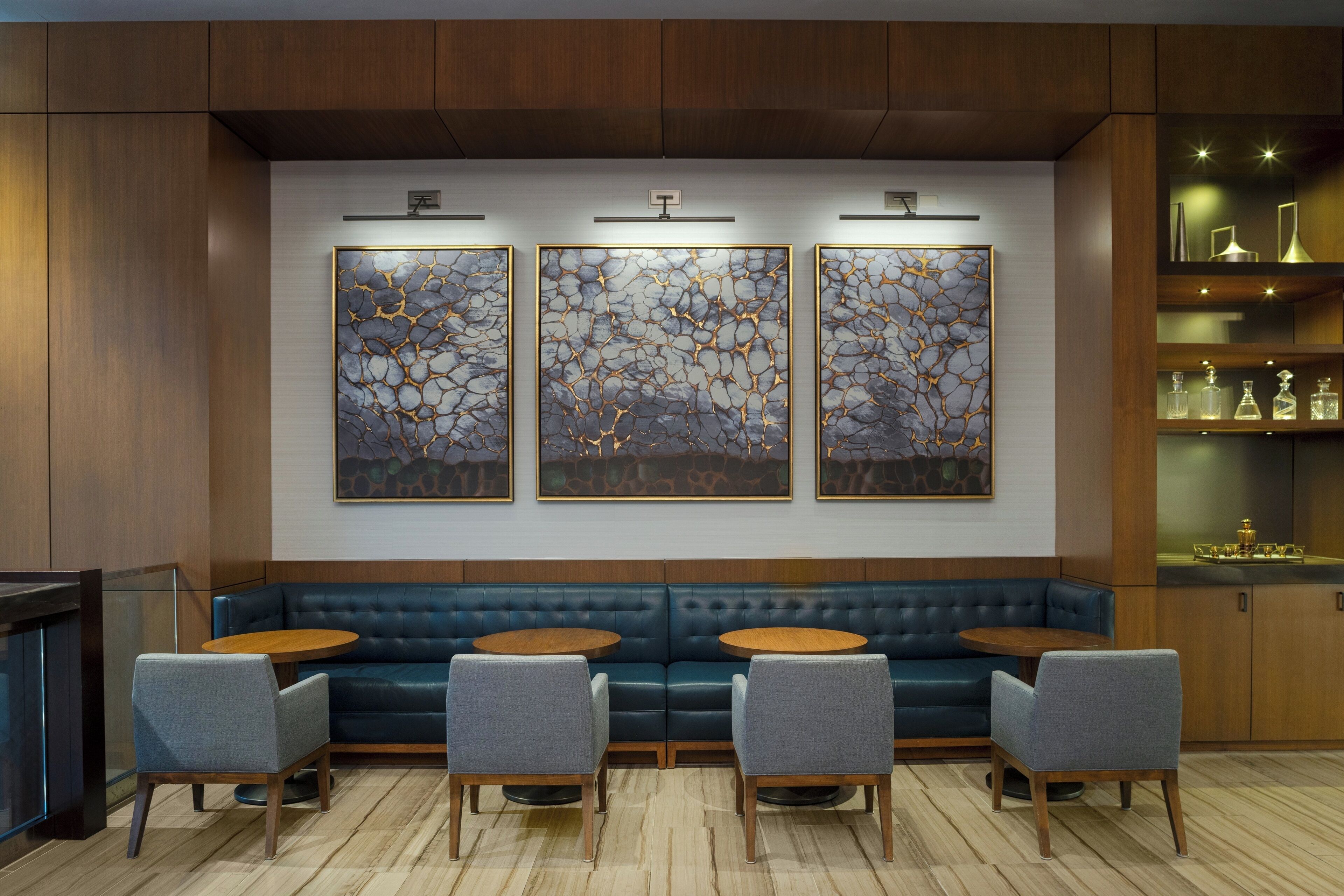 A modern interior with a blue leather sofa, four grey armchairs, and a wooden coffee table. There are three abstract paintings on the wall and a shelf with decorative items.