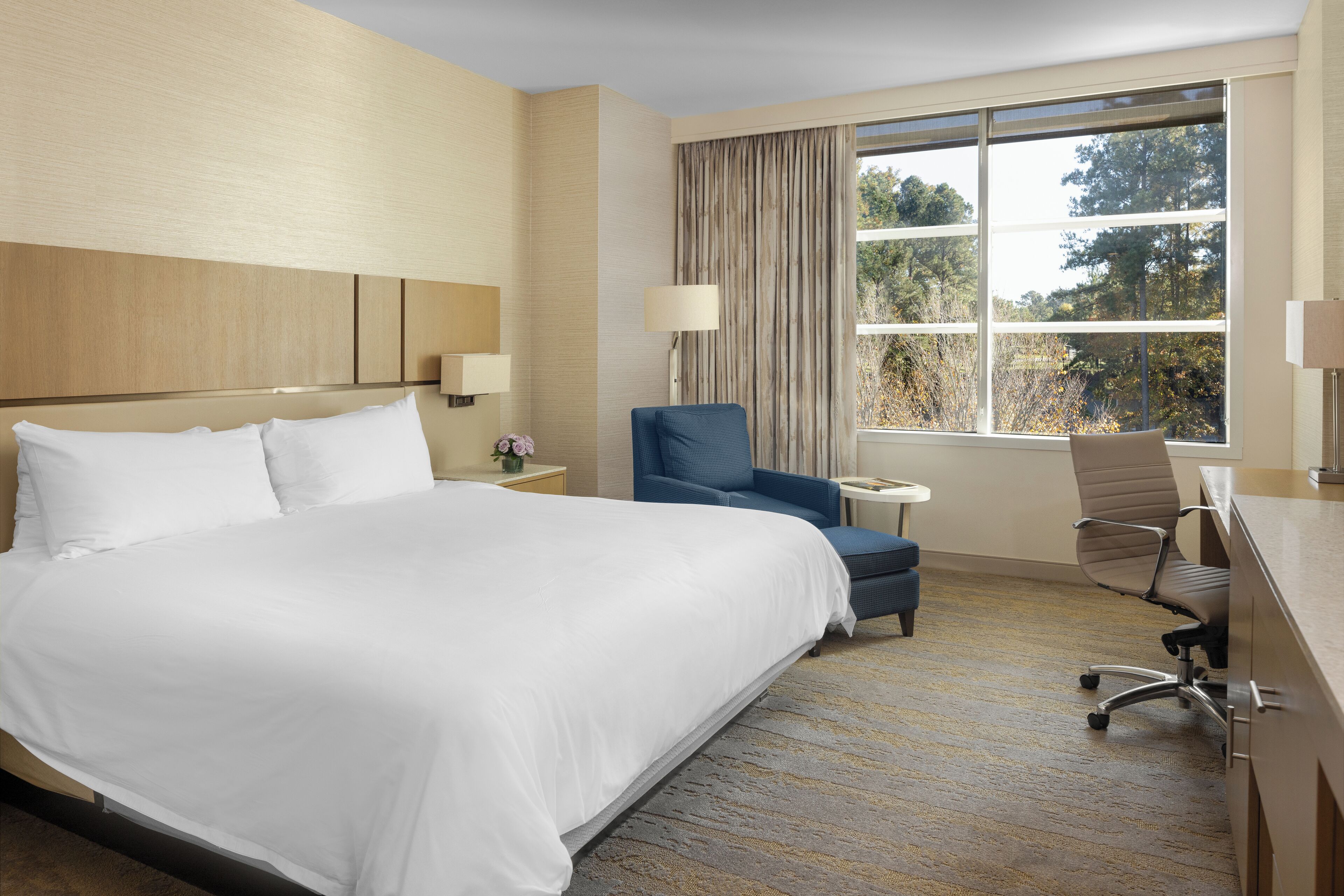 A hotel room with a large bed, a desk, a chair, a lamp, and a window with curtains.