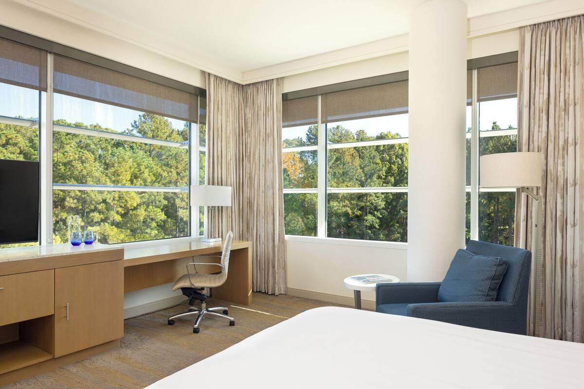 A hotel room with a large bed, a desk with a chair, a small round table, and a sofa. There are large windows with curtains, and a view of trees outside.