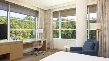 A hotel room with a large bed, a desk with a chair, a small round table, and a sofa. There are large windows with curtains, and a view of trees outside.