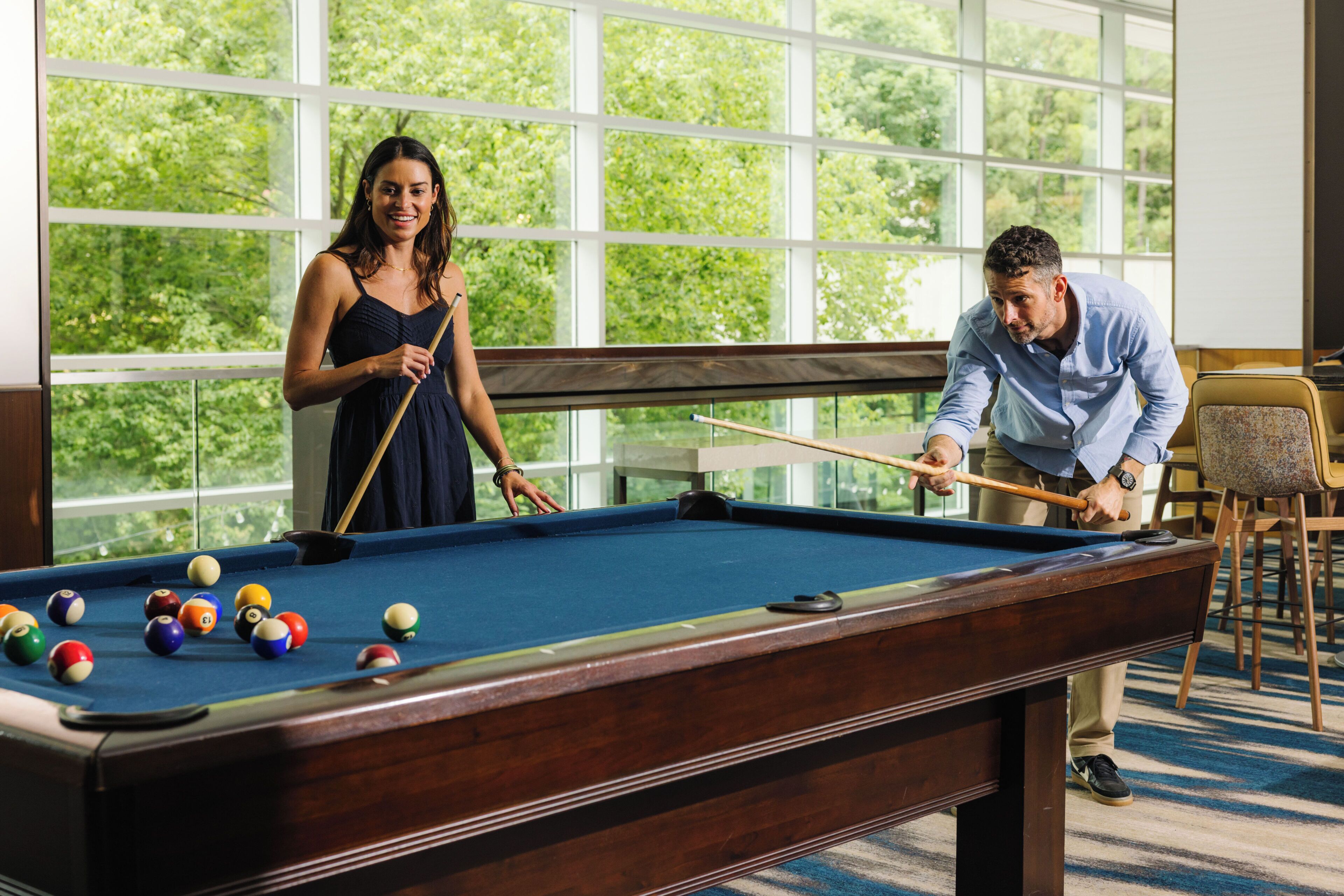 Une table de billard avec des billes, une femme et un homme qui jouent, et une vue sur la verdure à travers de grandes fenêtres.