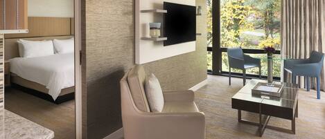 A modern hotel room with a bed, a sofa, a TV mounted on the wall, and a view of the outdoors.