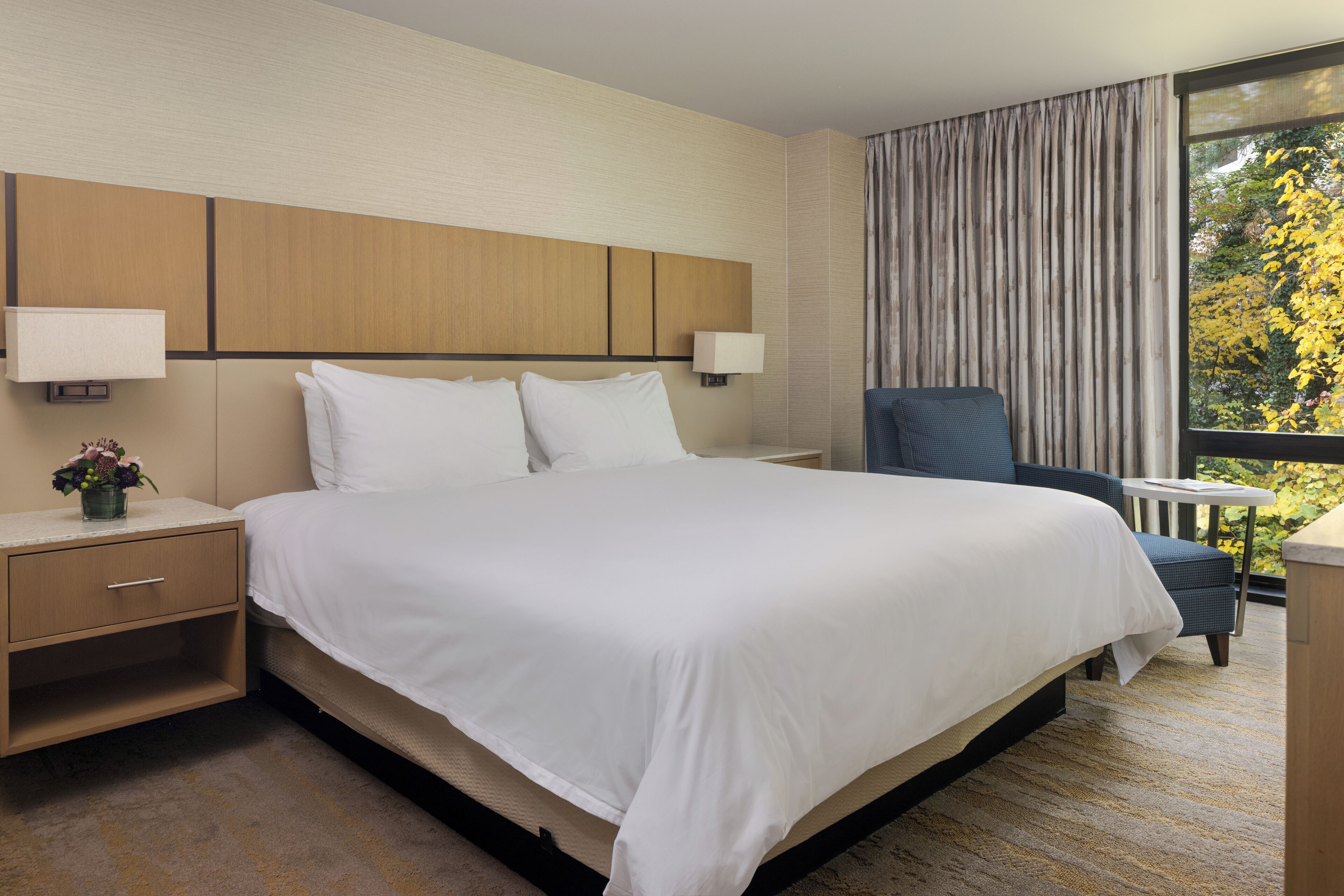 Een hotelkamer met een groot bed, twee bedlampen, een nachtkastje met een bloevas, een stoel en uitzicht op bomen door een raam.
