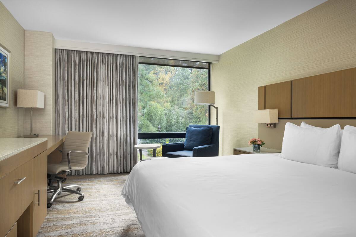 A hotel room with a large bed, a desk with a chair, a small round table, and a window with curtains.