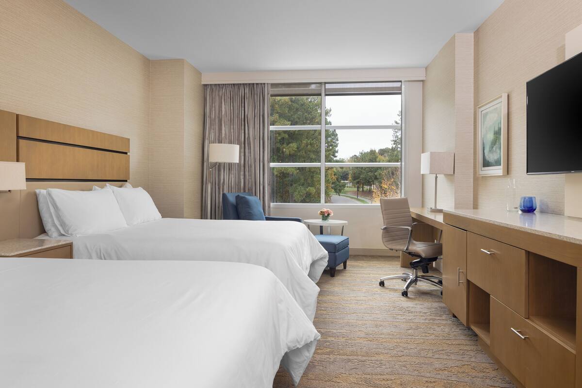 A hotel room with two beds, a desk, a chair, and a TV.