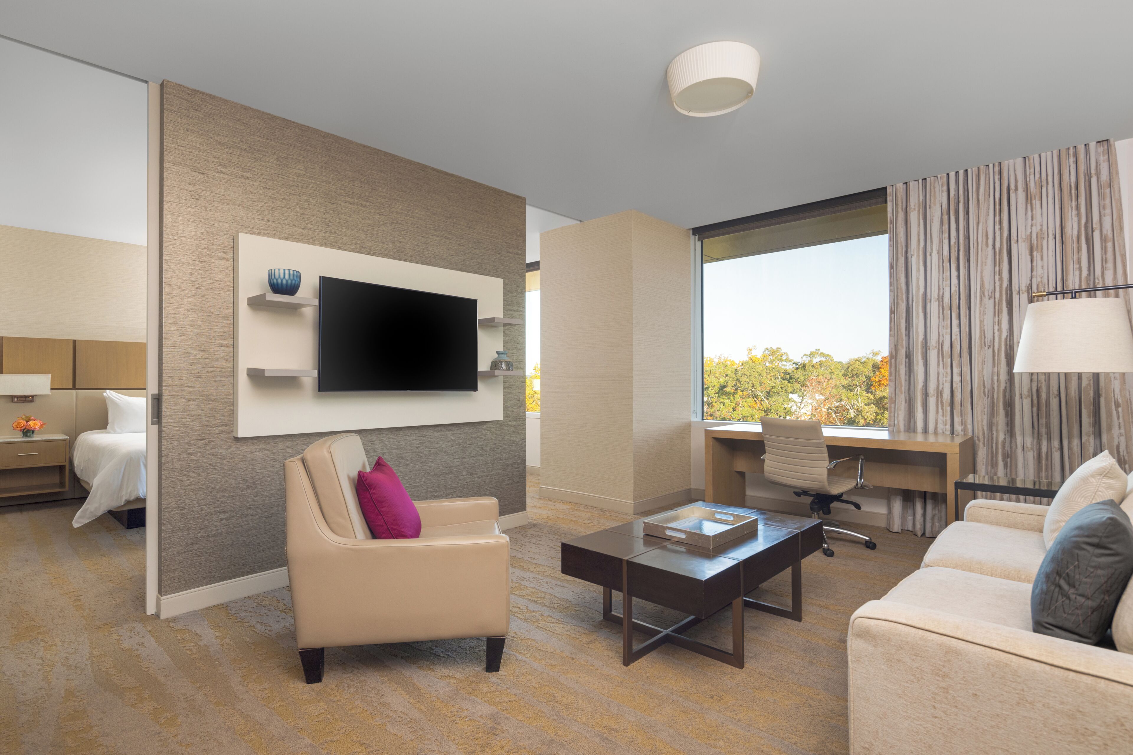 A modern hotel room with a flat-screen TV mounted on the wall, a desk with a chair, a sofa, and a coffee table.
