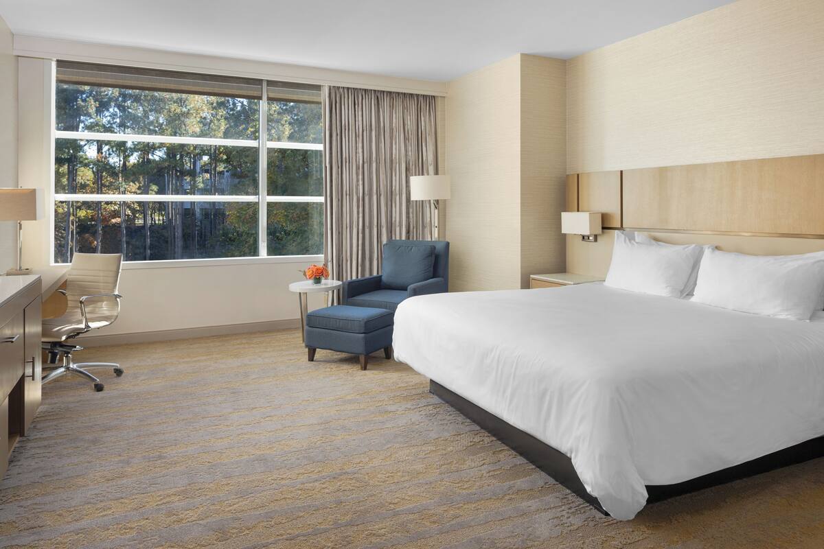 A hotel room with a large bed, a desk with a chair, a small table with a lamp, and a view of trees through the window.