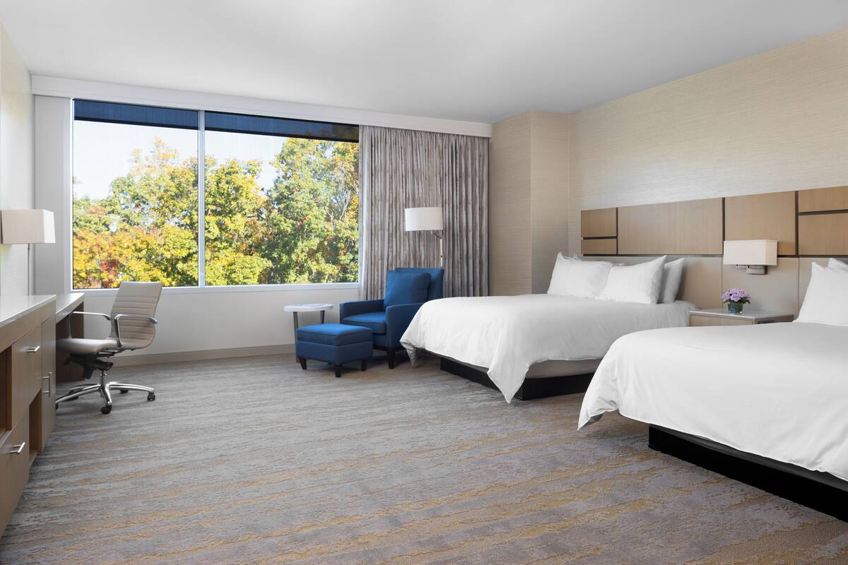 A hotel room with two beds, a desk, a chair, and a large window with a view of trees.