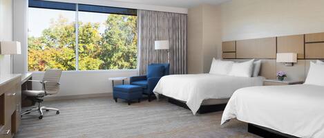 A hotel room with two beds, a desk, a chair, and a large window with a view of trees.