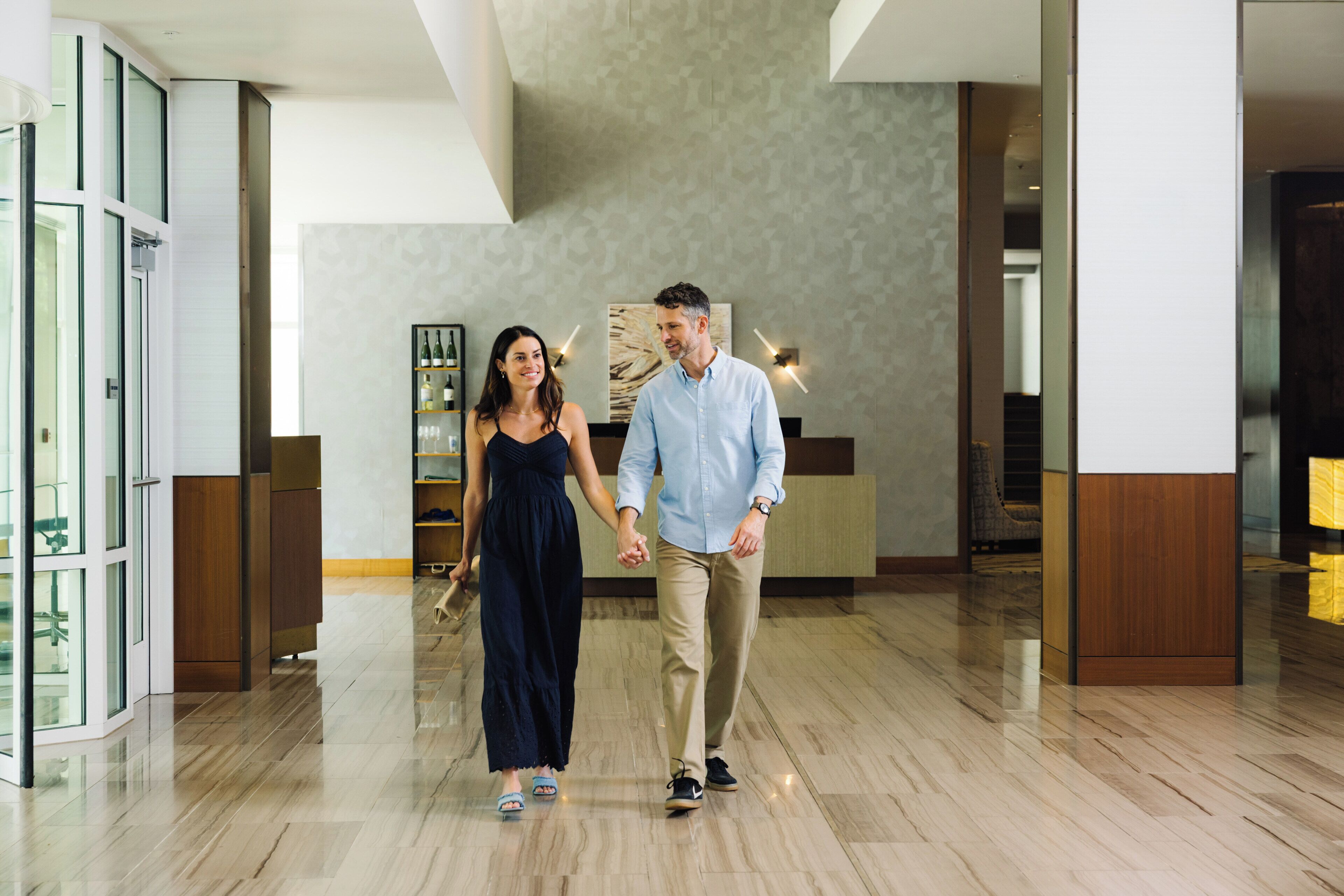 A couple walking through a hotel lobby with a modern design, including a reception desk and a wine display.