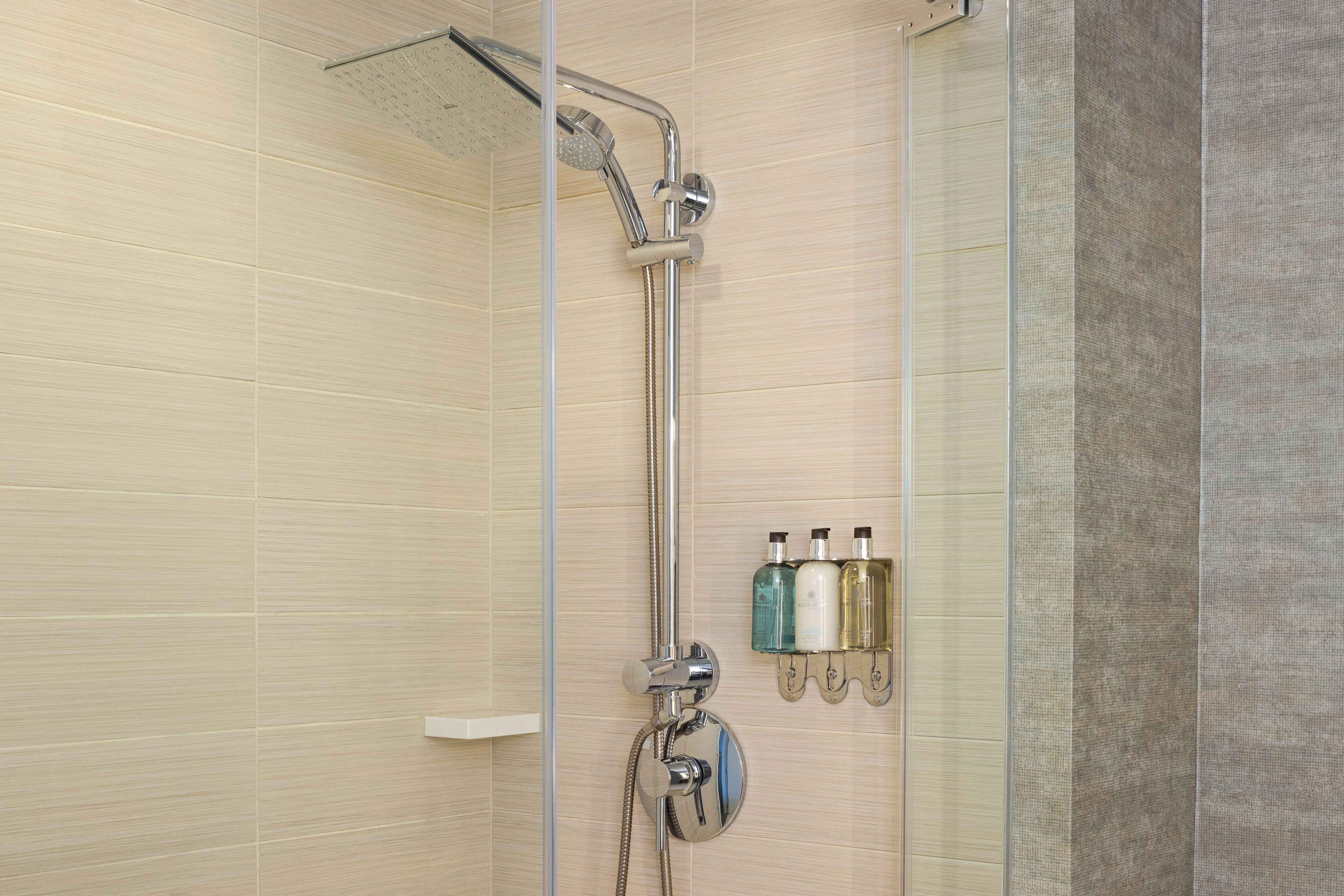 A shower with a handheld showerhead, a wall-mounted faucet, and a glass shower caddy with bottles.