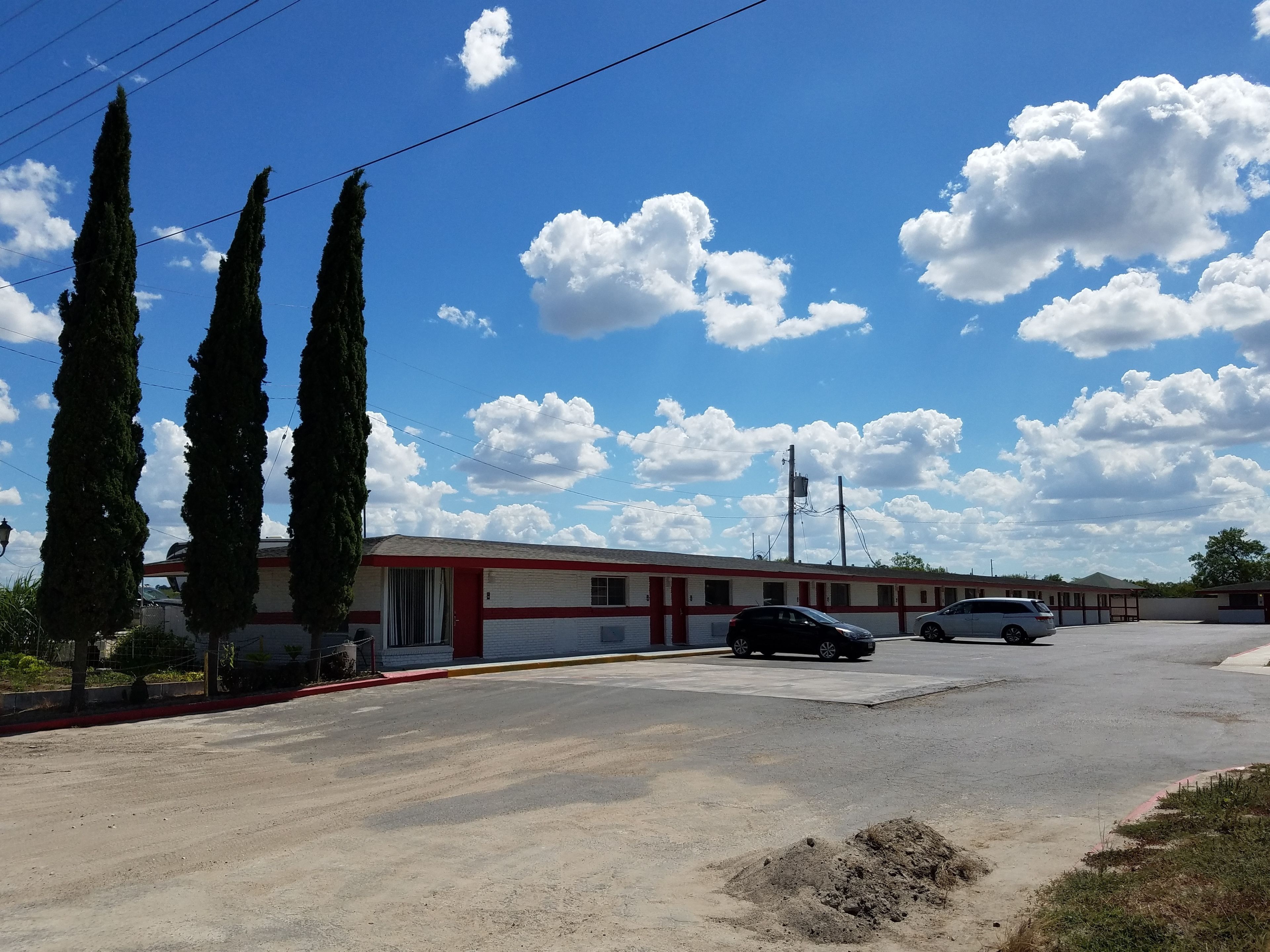 A motel with a red and white facade, two parked cars, and a clear blue sky with clouds.