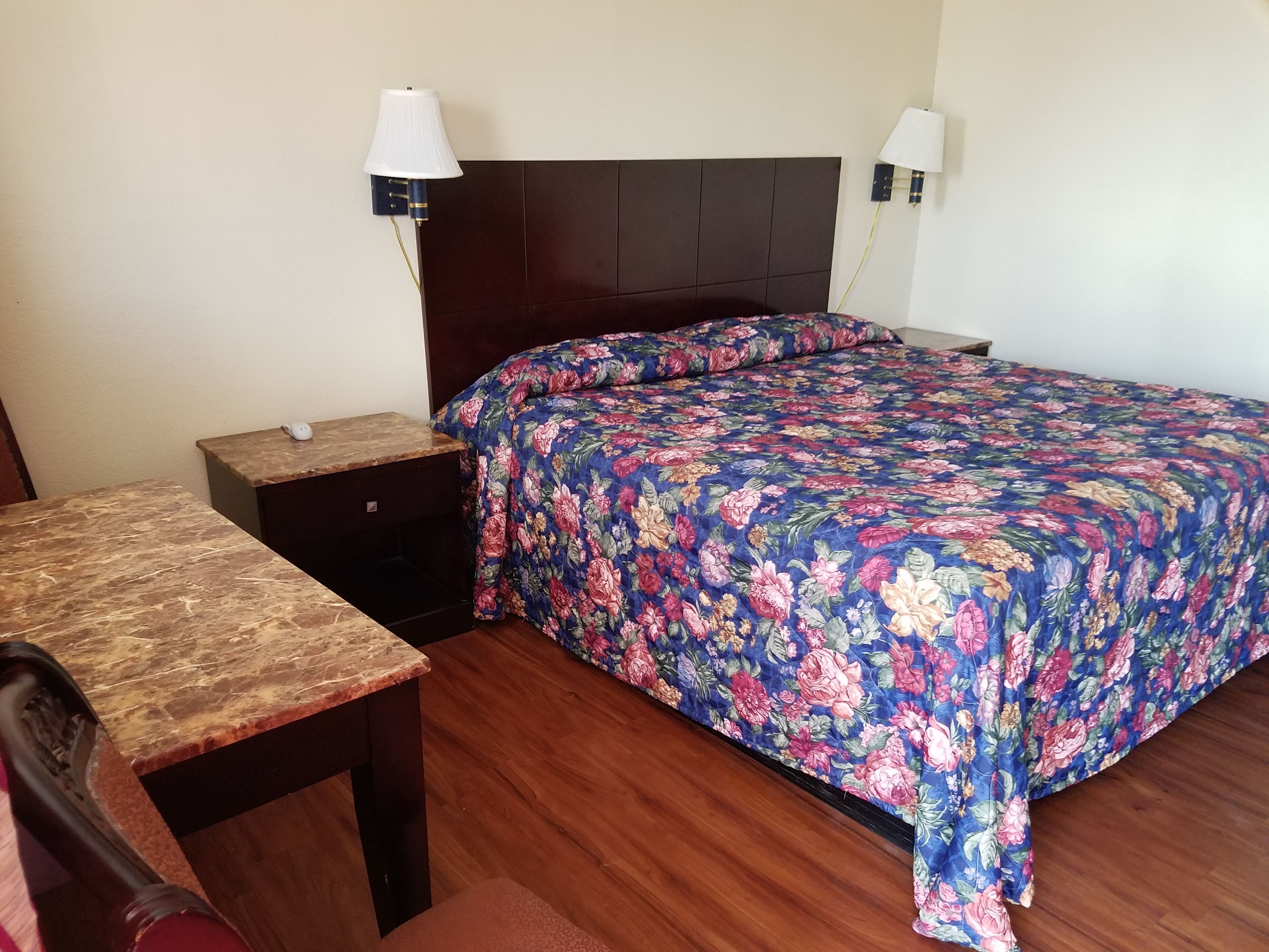 Una camera d'albergo con un letto di legno, un comodino, una scrivania e due lampade a parete.