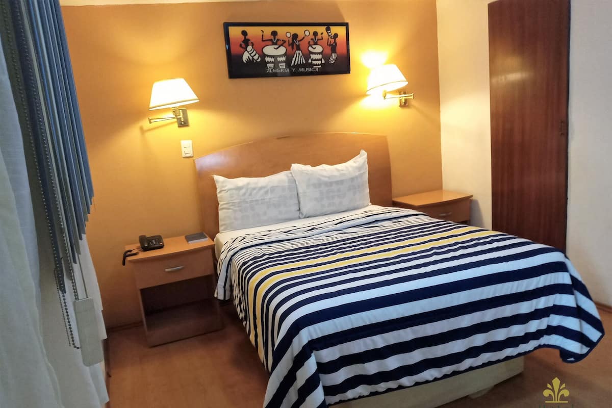A hotel room with a bed, two bedside lamps, a nightstand, a wall-mounted picture, and a wooden door.