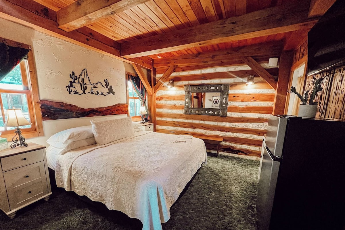 A cozy log cabin bedroom with a large bed, a nightstand, a dresser, a mirror, and a refrigerator.