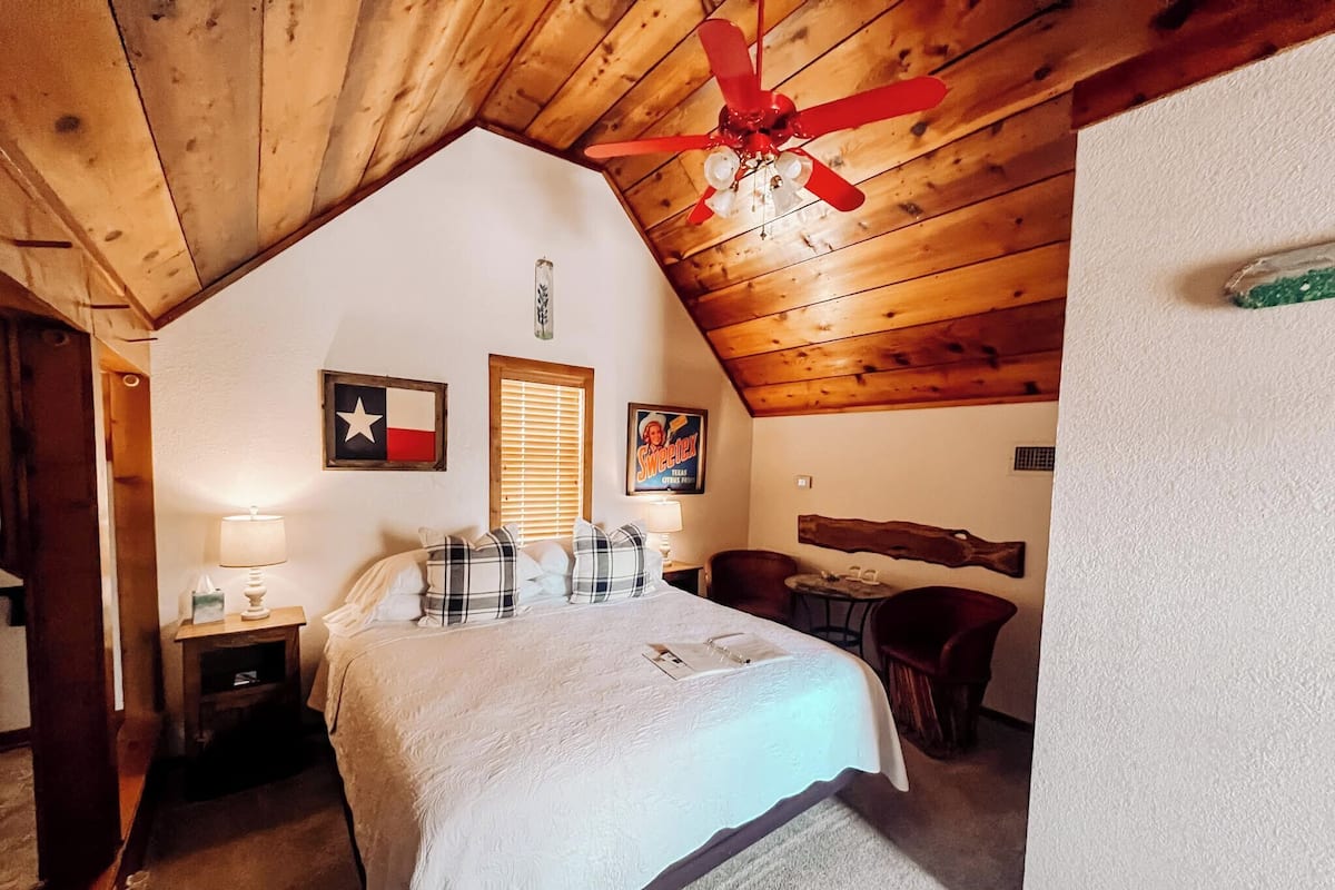 A cozy room with a bed, a wooden ceiling, a ceiling fan, a window with blinds, and a Texas flag on the wall.