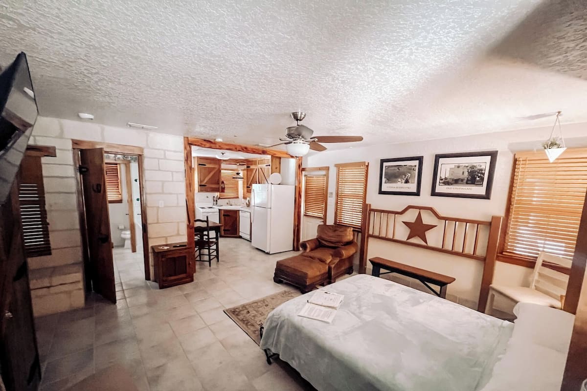 A bedroom with a bed, a desk, a chair, a ceiling fan, and a kitchenette in the background.