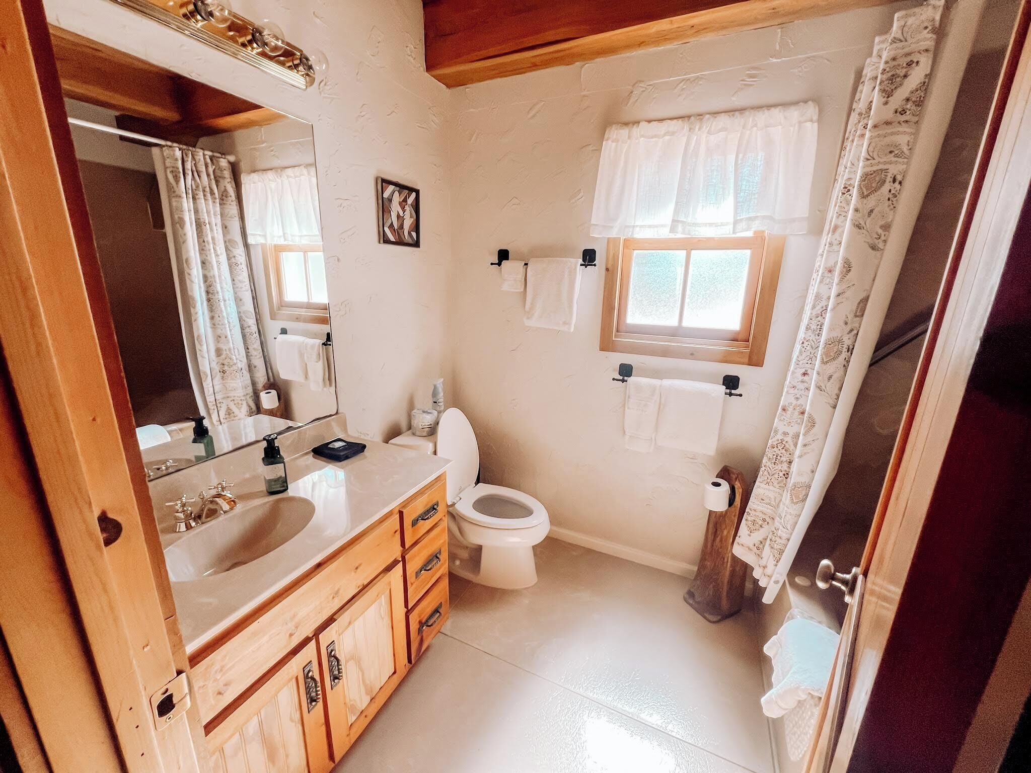 A bathroom with a wooden vanity, a toilet, a sink, and a window with a curtain.