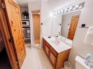 A bathroom with a wooden vanity, a large mirror with lights, a toilet, and a shower area. - Gruene Cottages (New Braunfels)