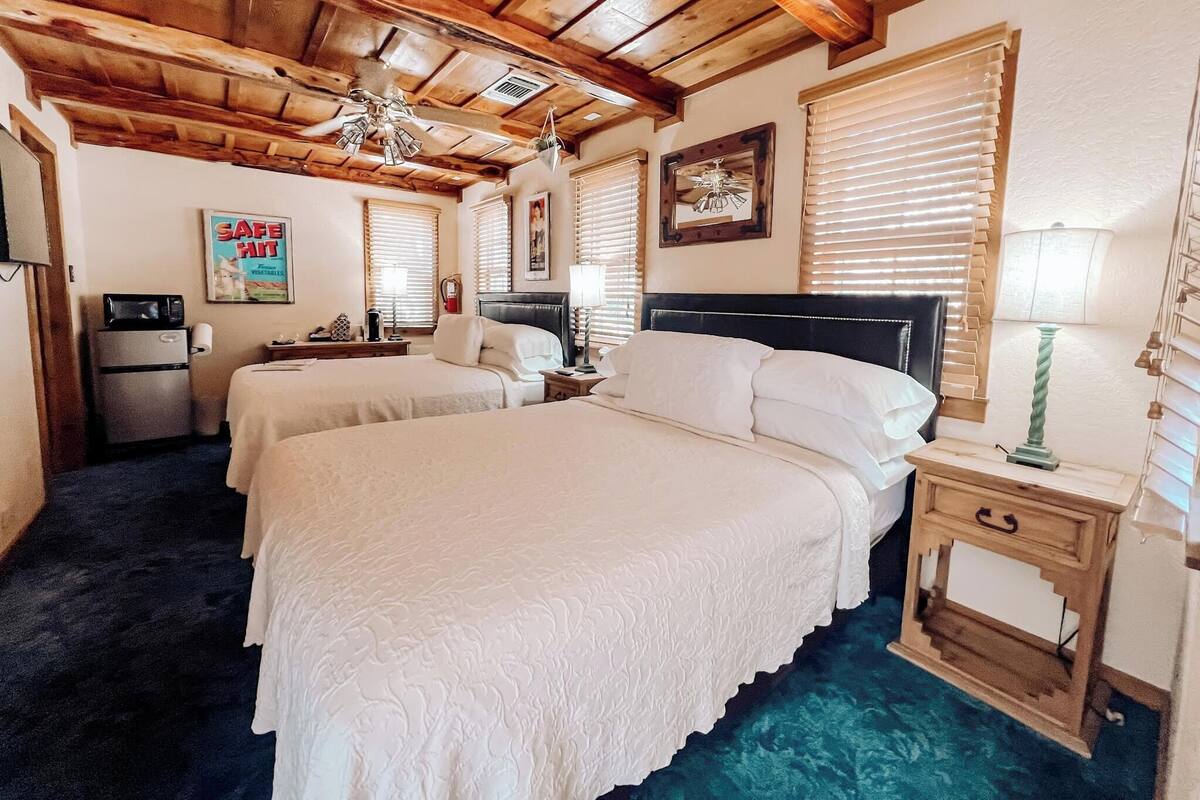 A hotel room with two beds, wooden ceiling beams, a small kitchenette, and a large window with blinds.