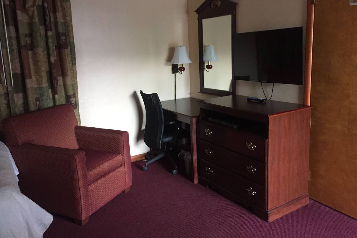 A hotel room with a red armchair, a desk with a mirror, a television, and a dresser.